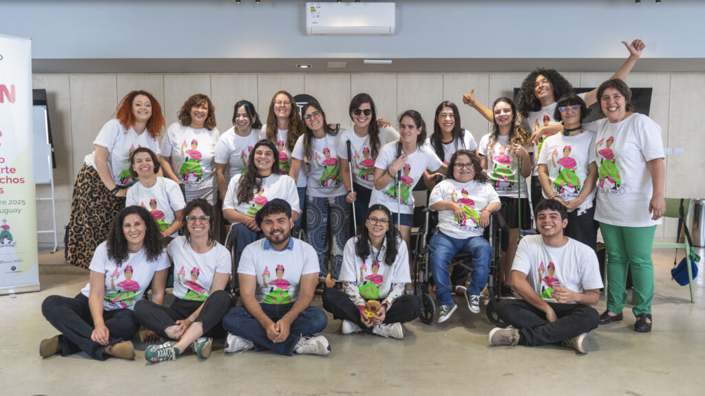 Foto grupal de más de veinte personas, varias de ellas en sillas de ruedas participantes del encuentro Fogón del Sur. Todas llevan remeras blancas con la ilustración colorida del logo de La Luz de Frida. Están sonriendo en un salón amplio y luminoso.