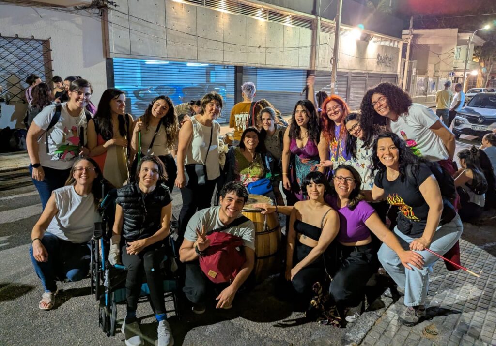 Un grupo grande de personas jóvenes y adultas posa sonriente en una calle iluminada de noche. Algunas están sentadas o en sillas de ruedas. Detrás se ven autos estacionados y un edificio con persianas metálicas.