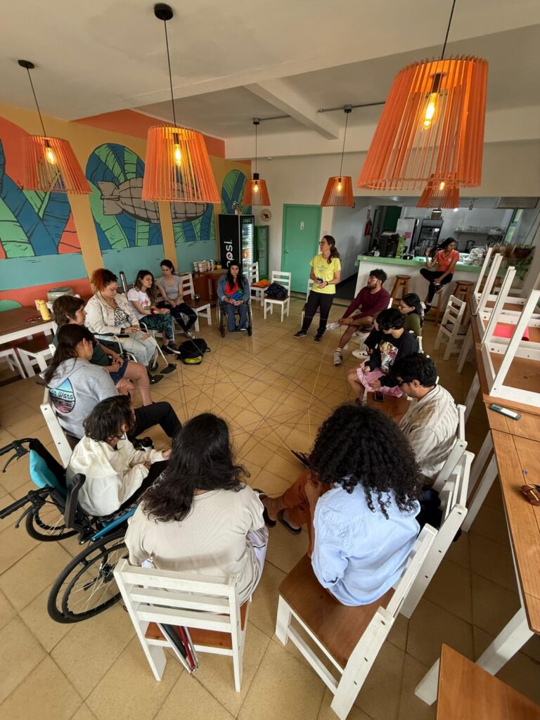 Un grupo de personas sentadas en círculo en un salón con paredes pintadas con hojas grandes y lámparas colgantes naranjas. Algunas personas están en sillas de ruedas. En el centro, cuerdas unidas forman una red entre quienes participan.