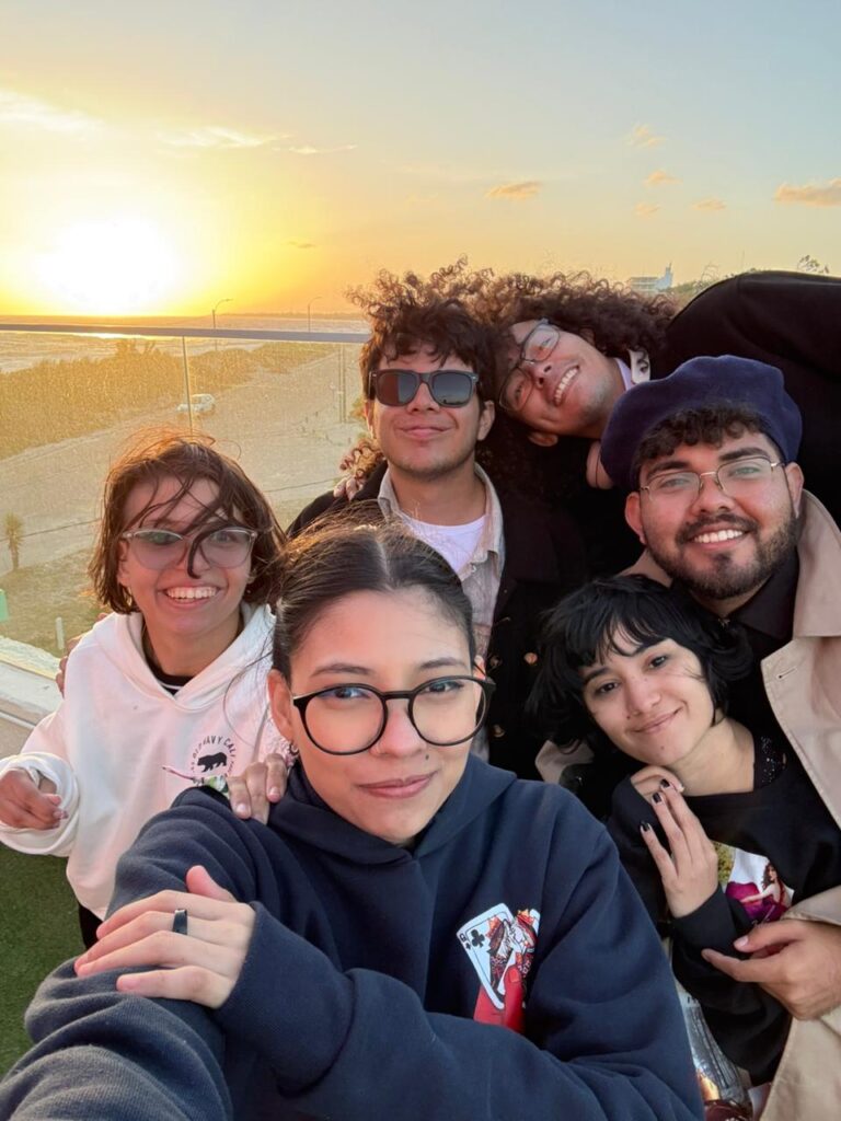 Seis jóvenes posan sonrientes para una selfie al aire libre, con el mar y el sol cayendo en el horizonte detrás de ellos. Llevan camperas y buzos, y el viento les mueve el pelo.