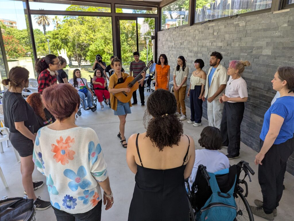 Un grupo de jóvenes y adultos, algunas personas en sillas de ruedas, forman un círculo en un espacio vidriado y luminoso. En el centro, una persona toca la guitarra mientras el resto observa o canta. Afuera se ve un parque con árboles.